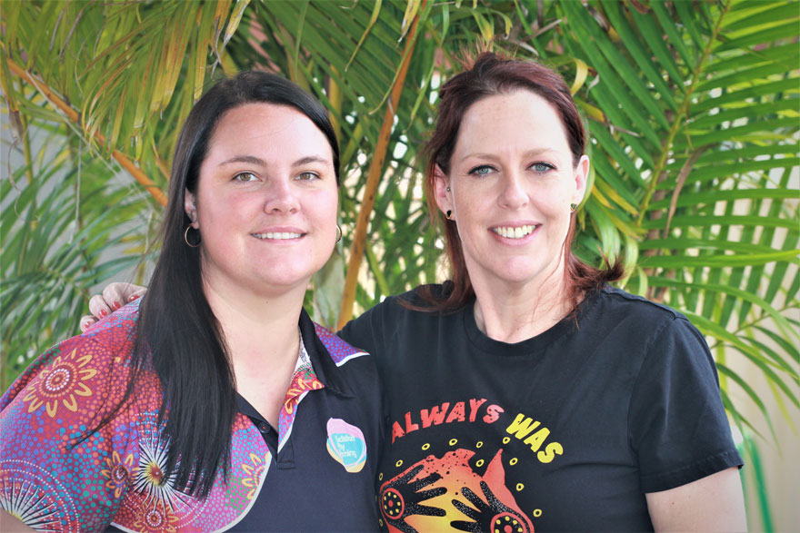 Two child care workers smiling and wearing colourful indigenous Australian t-shirts, with green, leafy plants in the background.