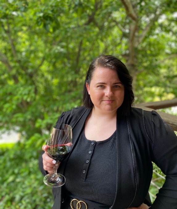 Image of a woman standing in front of greenery holding a glass of wine