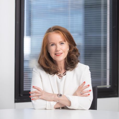 Head shot of Dr Lisa O'Brien AM with shoulder length red hair wearing a white blouse and a gold neck lace